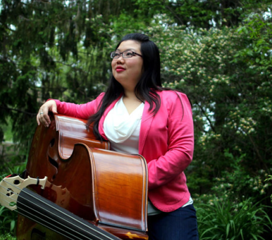 Woman in a pink blazer with glasses leans on a double bass outdoors among green trees.